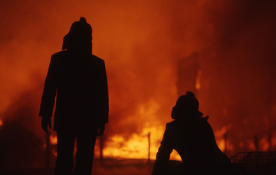 806139 Afbeelding van de silhouetten van twee brandweermannen tijdens het blussen van de in brand staande houten ...
